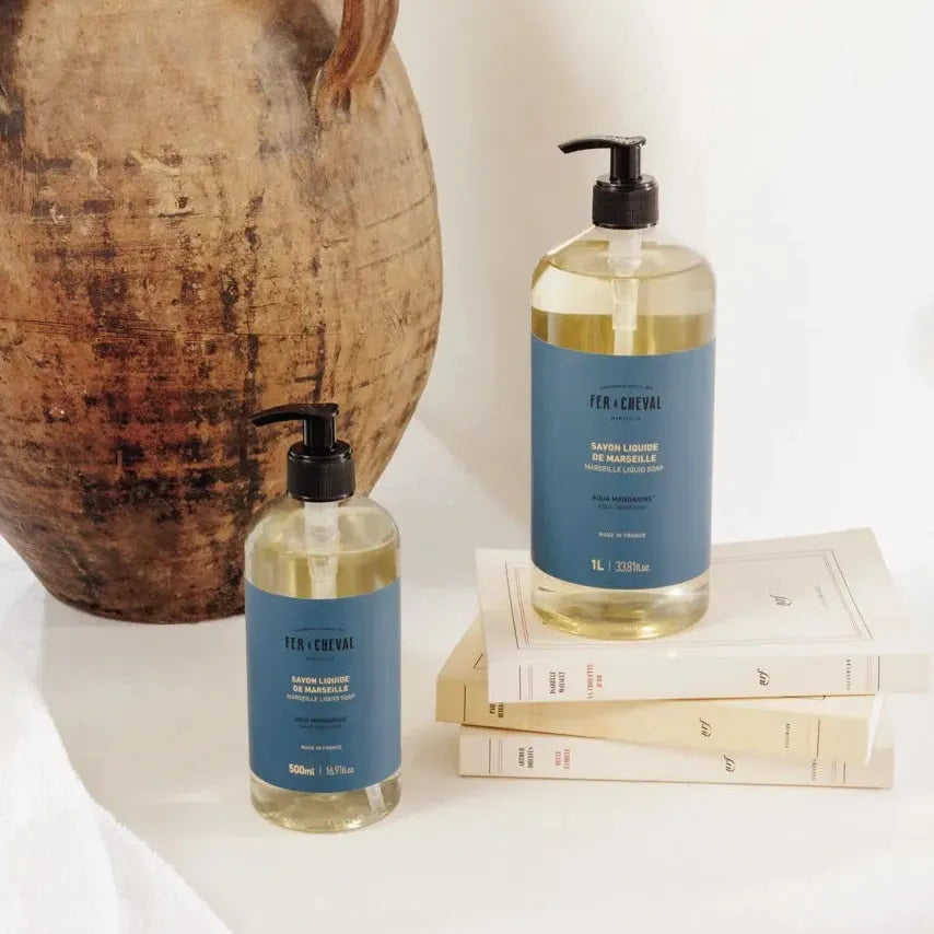 Two bottles of liquid soap with blue labels on a white surface next to a ceramic vase.