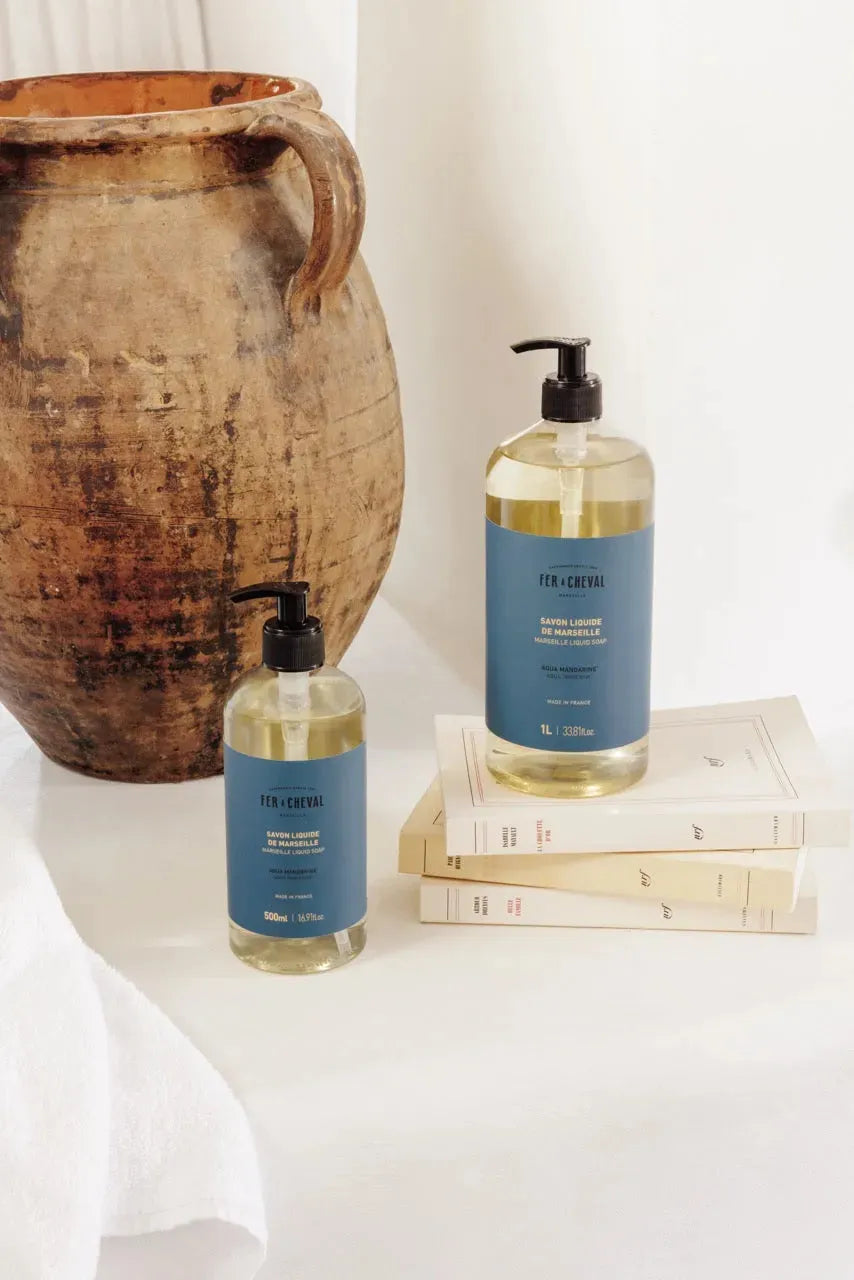 Two bottles of liquid soap with blue labels on a white surface next to a ceramic vase.