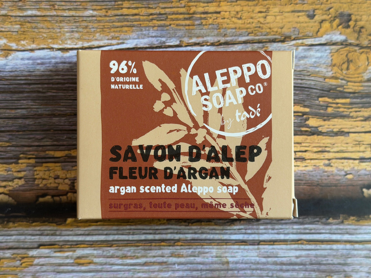 A rectangular box of Aileppo Soap Co. Savon D'Alep (Fleur D'argan) almond scented soap, featuring a brown and beige label, placed on a rustic, weathered wooden surface.