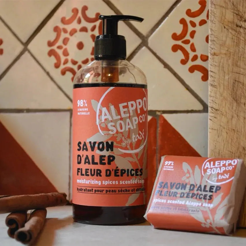 A Bottle of Liquid Aleppo Soap with a red label "Fleur d'spices, and a bar soap next to it with cinnamon sticks and a tile wall behind.