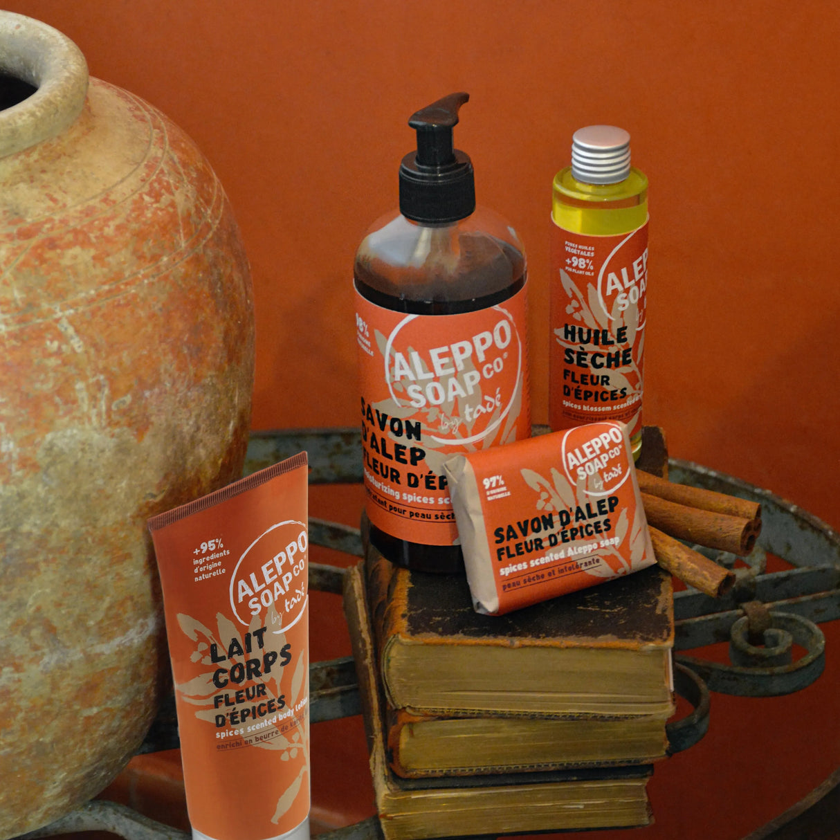A collection of Red labelled Aleppo Soap Co Products "flour d'epices' on a wrought iron table with a large terracotta pot and an orange painted wall