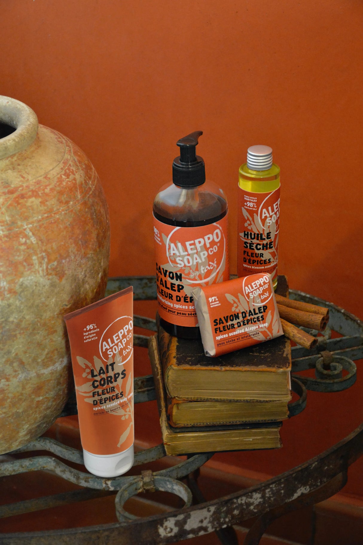 A collection of Red labelled Aleppo Soap Co Products "flour d'epices' on a wrought iron table with a large terracotta pot and an orange painted wall