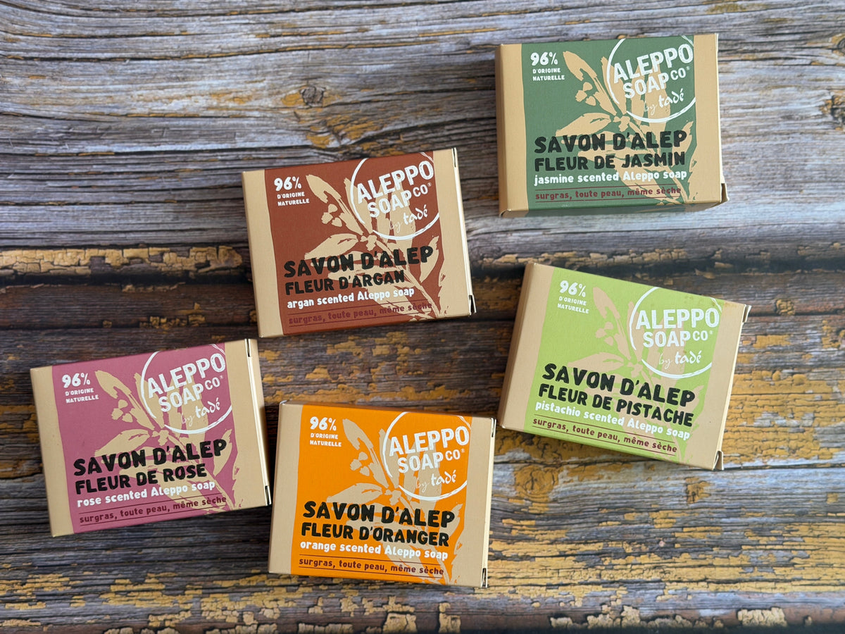 Five small rectangular soap boxes arranged on a rustic weathered wood surface. The boxes are in different colorways: pink rose, orange blossom, peach, pistachio green, and ash green, all branded ALEPO.