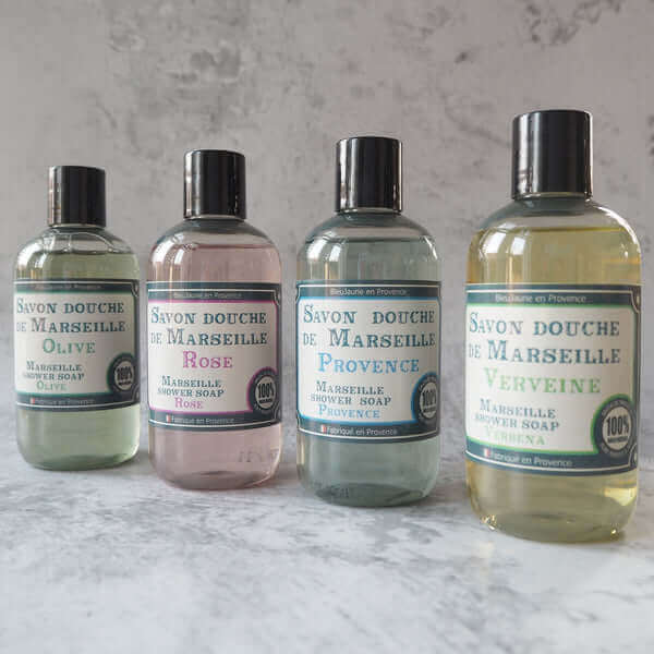 Four bottles of Savon douche de Marseille soap on a marble surface with a gray background. French Soaps UK