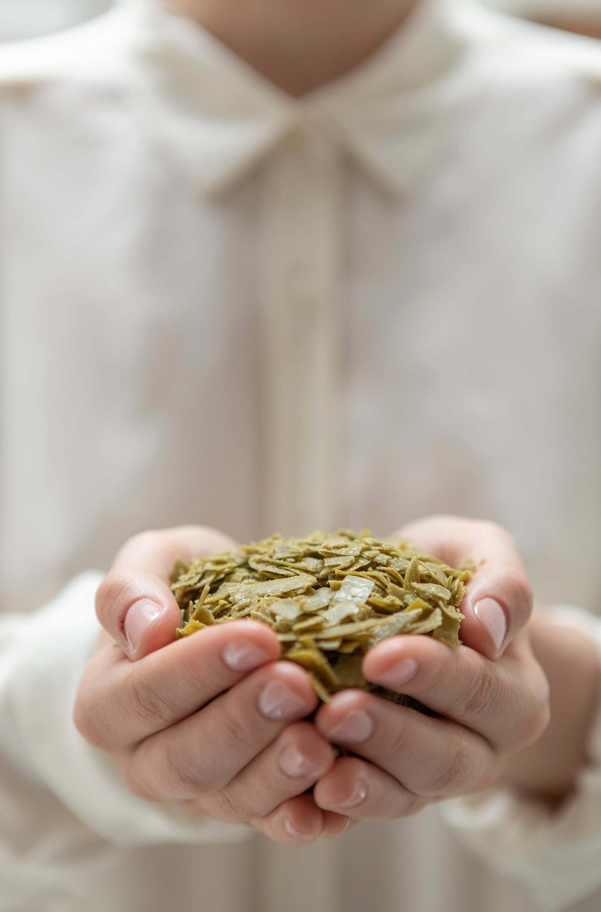 Person holding a small pile of green olive Marseille soap flakes in their hands against a blurred background. French Soaps UK