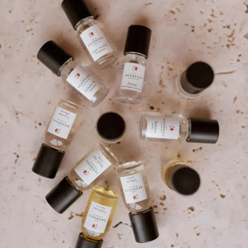 11 small glass bottles with black lids on a stone surface. The bottles are labeled Geodesis Fragrance Oil Concentrates with fragrances including Sakura, Verbena, Fig Tree, Agar Wood, Balsam Fir, Tuberose.