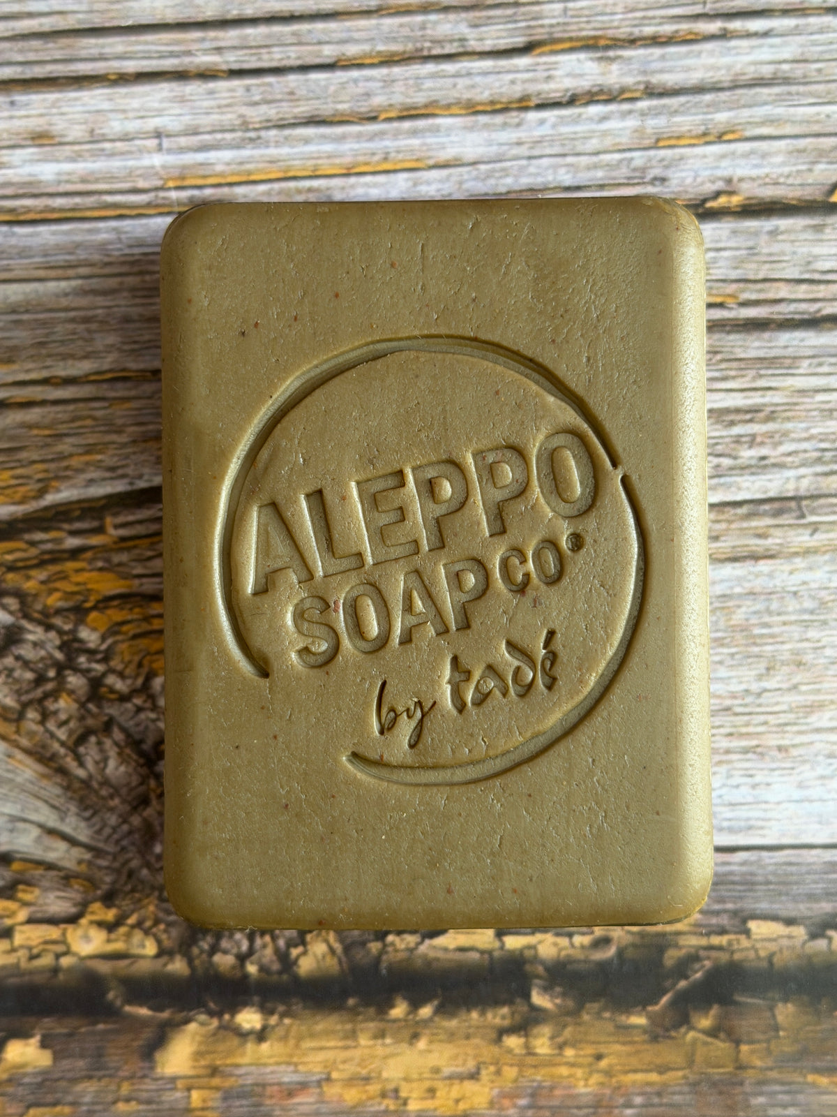 A rectangular bar of soap with rounded corners, embossed with the brand name 'ALEPPO SOAP CO. by tade', resting on a rustic wooden surface.