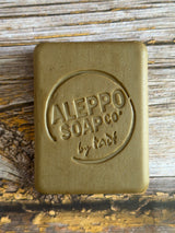 A rectangular bar of soap with rounded corners, embossed with the brand name 'ALEPPO SOAP CO. by tade', resting on a rustic wooden surface.