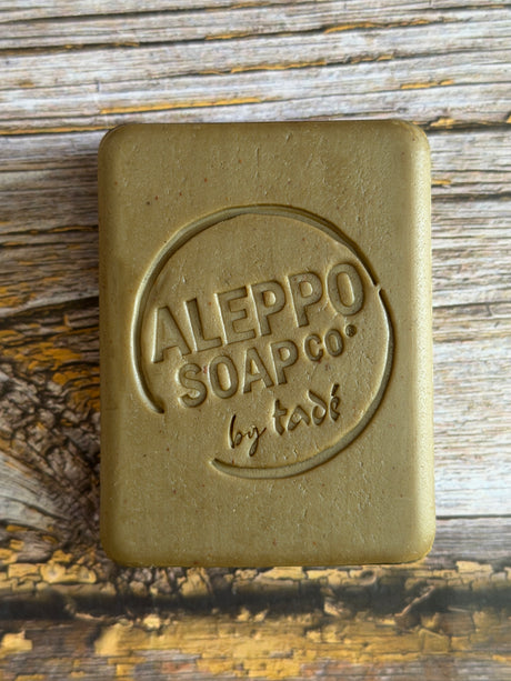 A rectangular bar of soap with rounded corners, embossed with the brand name 'ALEPPO SOAP CO. by tade', resting on a rustic wooden surface.
