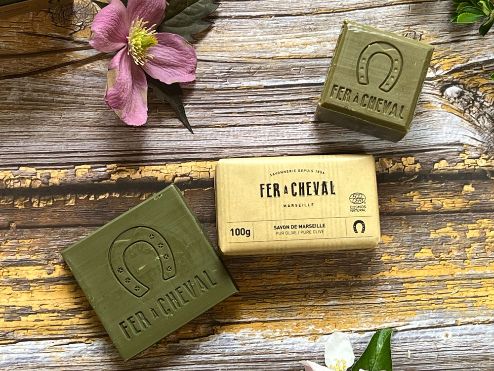 Two green soaps and a yellow wrapped bar  labeled 'Fer a Cheval' on a wooden surface with flowers. French Soaps UK