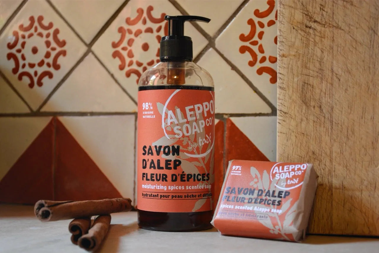 A Bottle of Liquid Aleppo Soap with a red label "Fleur d'spices, and a bar soap next to it with cinnamon sticks and a tile wall behind.