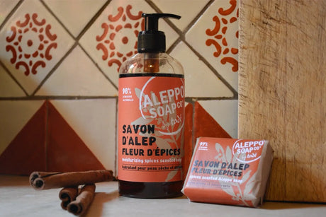 A Bottle of Liquid Aleppo Soap with a red label "Fleur d'spices, and a bar soap next to it with cinnamon sticks and a tile wall behind.