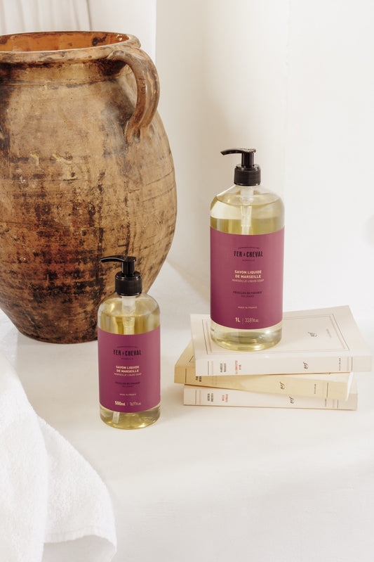 Two bottles of Fler & Co. soap on a white surface with a rustic vase and books in the background.