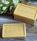 Two bars of Madeleine Marseille soap on a wooden surface with a sprig of lavender.