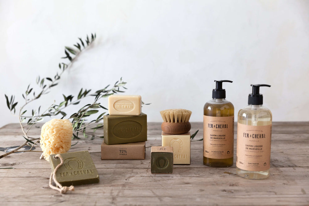 A Collection of Solid and Liquid Marseille soaps on a wooden table with an olive branch. French Soaps UK