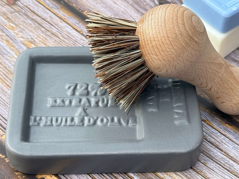 Wooden brush with bristles on a gray Square Soap dish labeled 'Huile d'Olive' on a wooden surface.