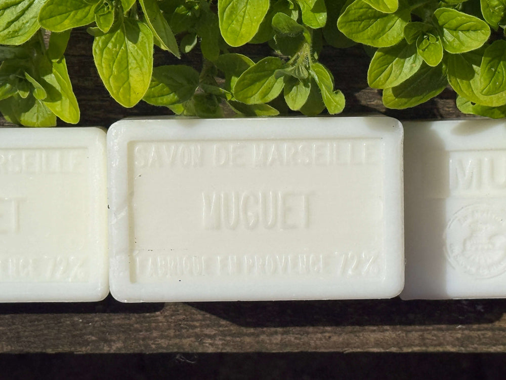 Three bars of soap with 'Muguet' Savon de Marseille branding on a wooden surface with green leaves in the background. Marseille Soap in the UK