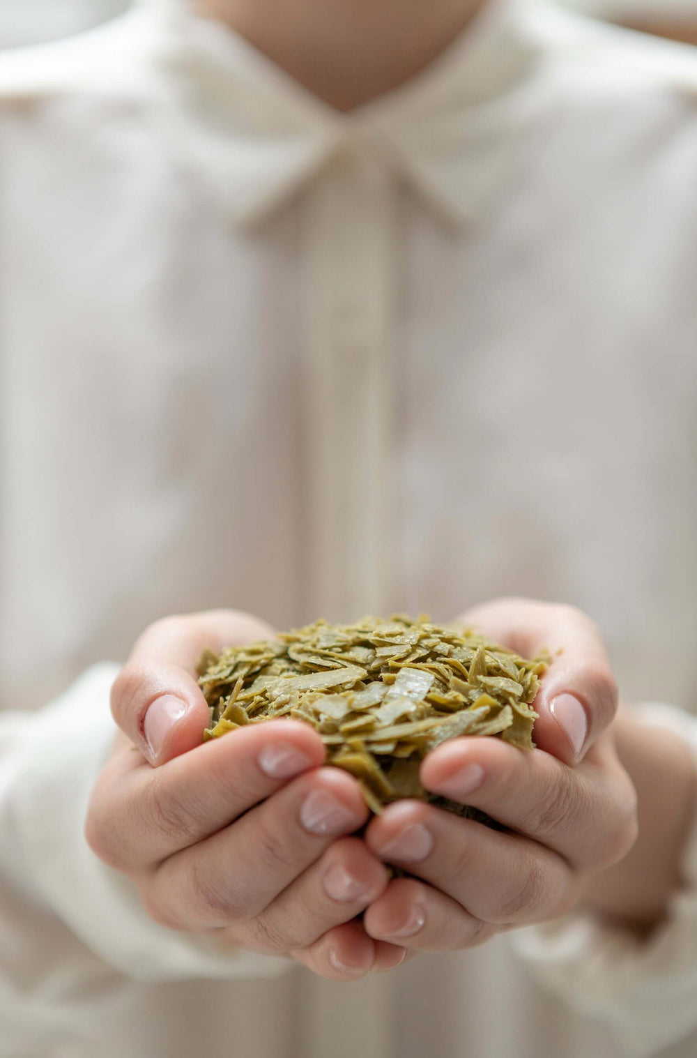 Person holding a small pile of green olive Marseille soap flakes in their hands against a blurred background. French Soaps UK 