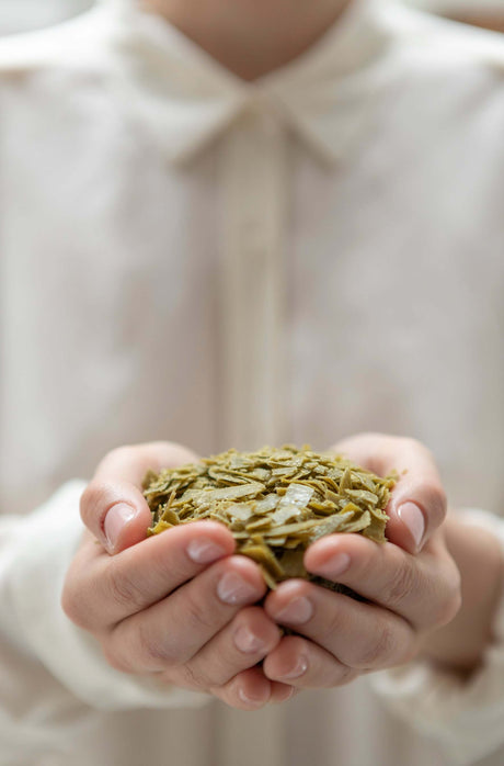 Person holding a small pile of green olive Marseille soap flakes in their hands against a blurred background. French Soaps UK 