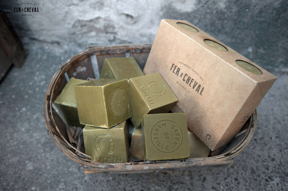 Green soap bars in a basket with a brown box labeled 'Fer a Cheval' on a gray surface.
