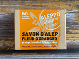 Aleppo Soap Co. Savon D'Alep orange scented Aleppo soap in a cardboard box with a bright orange label, shown on a rustic wooden surface.