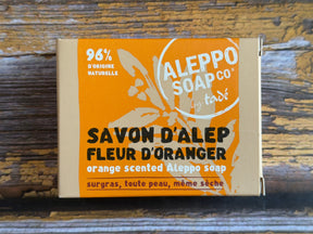 Aleppo Soap Co. Savon D'Alep orange scented Aleppo soap in a cardboard box with a bright orange label, shown on a rustic wooden surface.