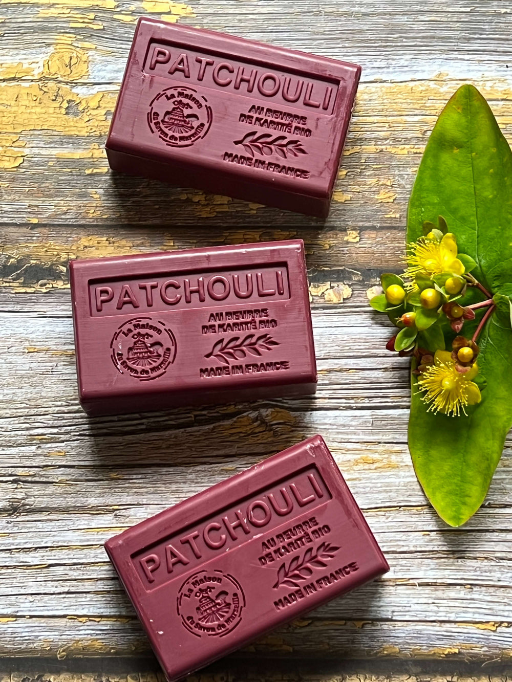 Three bars of Patchouli soap on a wooden surface with a leaf and flowers.