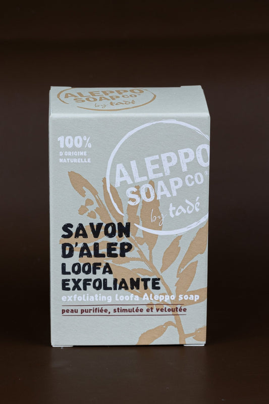A packaged bar of Aleppo Soap with Loofah Fibres on a dark background, with branding and product information displayed on the packaging.
