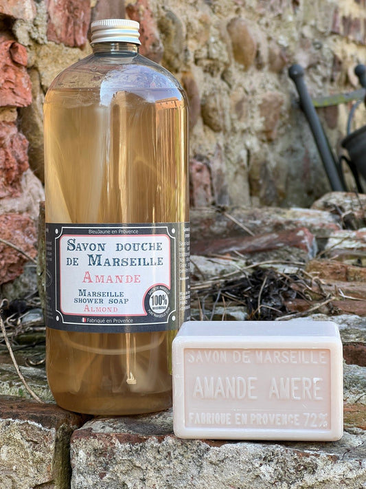 A clear plastic bottle of liquid soap labeled 'Savon doux de Marseille' alongside a bar of soap with 'AMANDE AURE' written on it, placed on a stone surface with a brick wall in the background.