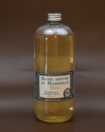 A clear plastic bottle containing yellow liquid soap, with a label that reads 'Savon douche de Marseille' and other text indicating it is a 1L bottle of honey-scented liquid soap.