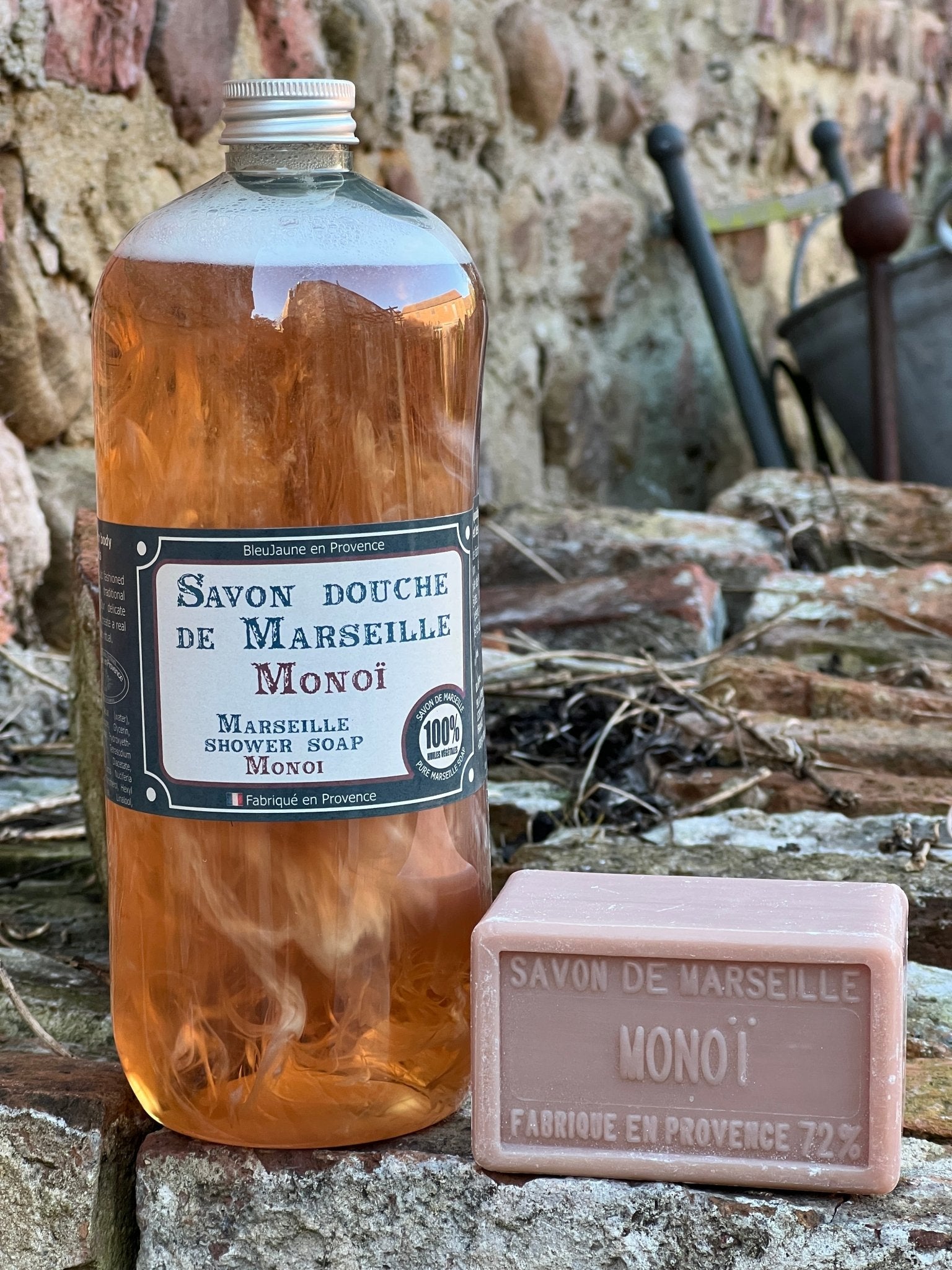 A clear bottle of liquid soap labeled 'Savon douche de Marseille' alongside a bar of soap with the same branding, set against a rustic stone background.