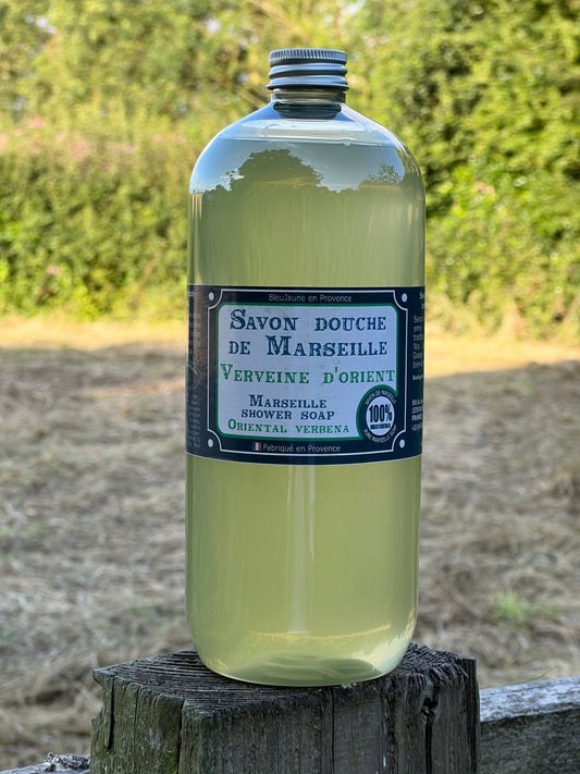 A clear plastic bottle of liquid soap labeled 'Savon de Marseille Oriental Verbena' placed on a wooden post outdoors.