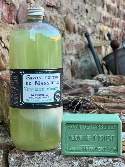 A clear bottle of Marseille liquid soap with a green label, alongside a green bar of soap, placed against a rustic background with a stone wall and old tools.