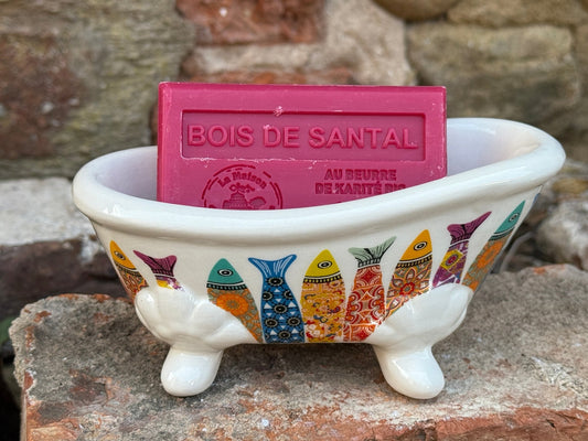Small bathtub-shaped soap dish with fish design holding a bar of soap against a stone background