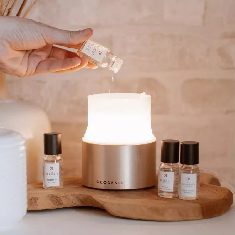 GEODESIS A large lit electric diffuser with fragrance oil being poured into it. The diffuser is on a shaped wooden board with three fragrance oil bottles situated either side of it.